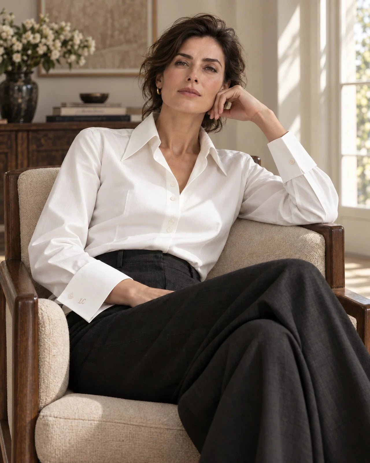 A woman in an ivory shirt with a generous collar and shaped cuffs, relaxed in an armchair, looking towards the camera, wearing charcoal trousers.